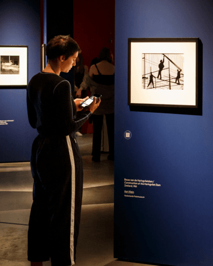 Vrouw staand voor een kunstwerk in museum met een telefoon in haar hand. Fotomuseum