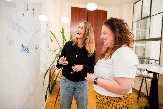 Twee vrouwen overleggen met elkaar, voor een whiteboard, lachen